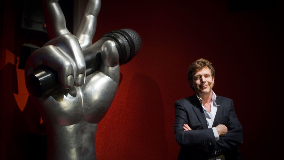 De Mol naast het iconische logo van "zijn" format, "The Voice"